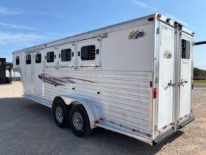 A silver Dream Coach 4 Horse trailer with four side windows and dual rear wheels is parked on gravel under a clear sky. The trailer features a rear door, side graphic decal, and ventilation panels. -Stillwater Trailer Sales