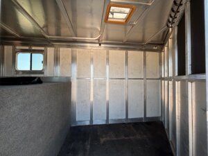 The image shows the interior of an empty Dream Coach 4 Horse Trailer with a window on the left, a carpeted partition, a rubber floor, and hooks on the upper right wall. There is a skylight on the ceiling. -Stillwater Trailer Sales