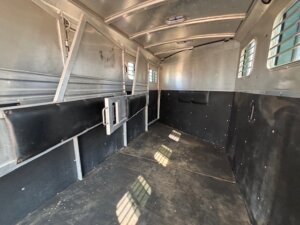 Interior of an empty Dream Coach 4 Horse trailer with partition dividers, barred windows, and a rubberized floor. Sunlight streams in through the windows, casting patterns across the spacious interior. -Stillwater Trailer Sales