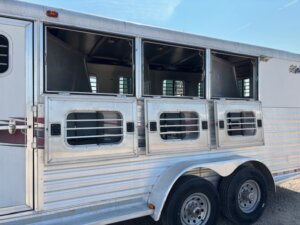 A silver Dream Coach Horse Trailer with three open windows showing barred doors. The 4 Horse trailer features two axles with four tires and horizontal stripes along the side, parked on gravel under a clear sky. -Stillwater Trailer Sales