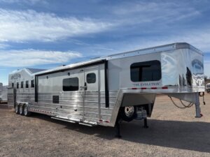 A large, silver Bloomer Signature 5 Horse gooseneck trailer named The Evolution is parked on a gravel lot under a clear blue sky. Its multiple windows and polished metal surfaces reflect the surroundings beautifully. -Stillwater Trailer Sales