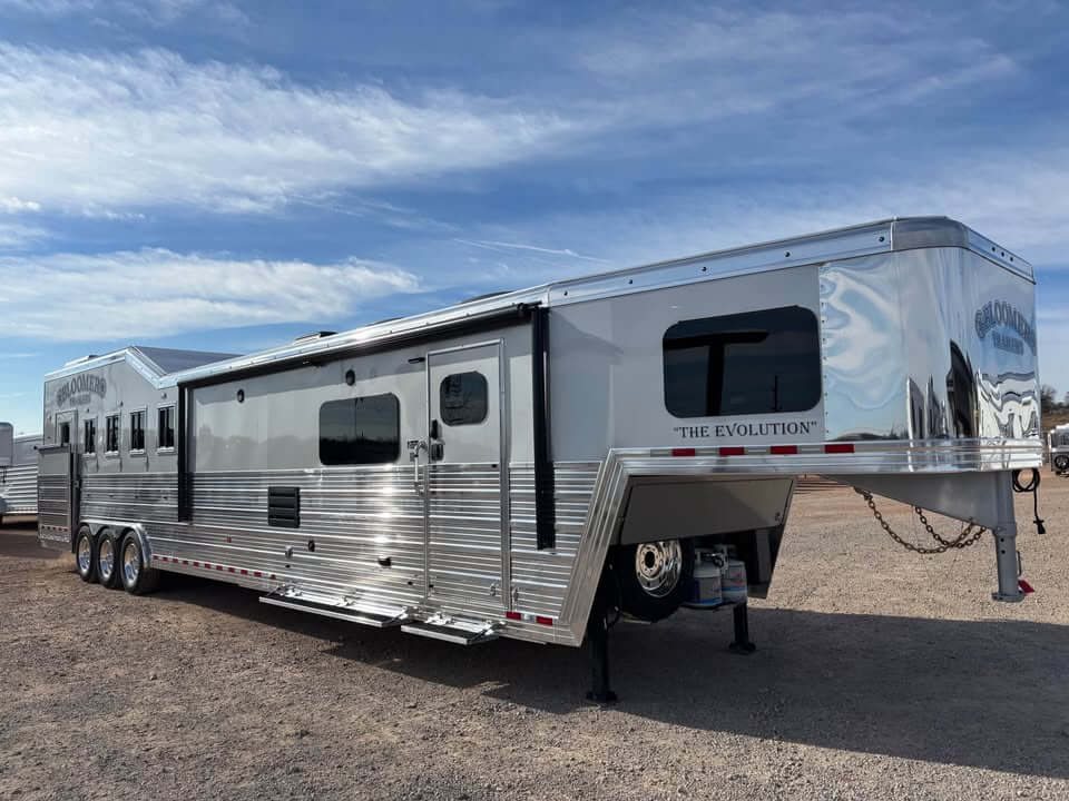 A large, shiny silver Bloomer horse trailer with multiple windows and several axles is parked on a gravel lot under a blue sky. The words “THE EVOLUTION” are visible on the front side of this impressive 5 horse trailer. -Stillwater Trailer Sales