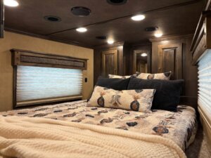 A neatly made bed with a Signature patterned comforter and pillows is in a cozy bedroom with wood cabinets, a window with closed blinds, and soft overhead lighting. -Stillwater Trailer Sales