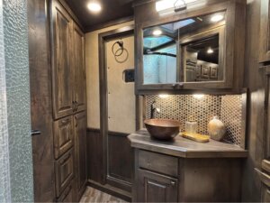 A bathroom with Signature dark wood cabinets, a vessel sink, a round mirror, a hexagonal tile backsplash, under-cabinet lighting, and decorative items on the counter. A frosted glass shower is partially visible on the left. -Stillwater Trailer Sales