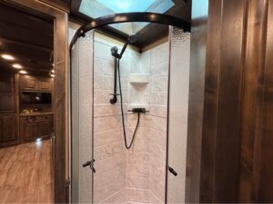 A corner shower with frosted glass doors, a black showerhead, and a tiled interior sits inside this Bloomer 5 Horse 17' Signature RV, complemented by wood-tone walls and flooring in the background. -Stillwater Trailer Sales