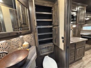 A Signature Bloomer camper bathroom features wood cabinets, an open tall storage unit with shelves, a vessel sink, tile backsplash, toilet, and a view into the kitchen and sleeping area. -Stillwater Trailer Sales