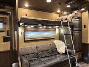 A compact living space features a gray couch below a lofted bed with a pillow and blanket, accessed by a metal ladder. Signature overhead lighting and wood paneled walls complete the cozy interior. -Stillwater Trailer Sales