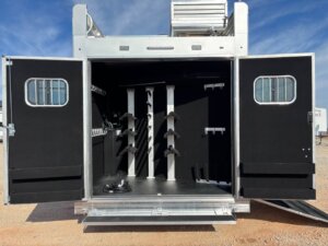 The image shows the open rear of a silver Bloomer trailer with black interior walls, equipped with racks and holders for storage, a coiled cable on the floor, and two open doors revealing the organized space inside this Signature 5 Horse model. -Stillwater Trailer Sales
