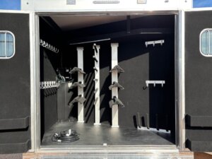 The image shows the open rear compartment of a 17' Signature Bloomer horse trailer with saddle racks, bridle hooks, and storage areas. The interior is lined with dark material, and a coiled cable is on the floor. -Stillwater Trailer Sales
