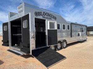 A large silver Bloomer 5 Horse trailer with multiple doors and ramps open, revealing Signature storage compartments and interiors, parked on gravel under a clear blue sky. -Stillwater Trailer Sales