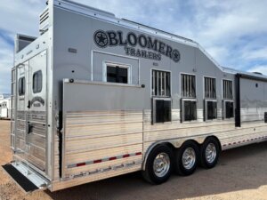 A large, silver Bloomer Signature 5 Horse trailer with three axles is parked on gravel. The trailer features multiple barred windows and a rear door, with the Bloomer Trailers logo visible on both the side and door. -Stillwater Trailer Sales