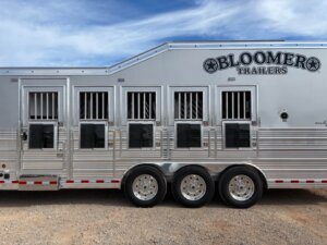 A silver Signature Bloomer 5 Horse trailer with six barred windows and three axles is parked on gravel under a blue sky with scattered clouds. BLOOMER TRAILERS is written on the upper right side. -Stillwater Trailer Sales