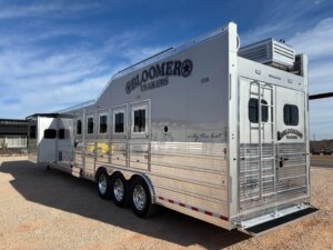 A large silver Bloomer 5 Horse trailer with multiple windows, three axles, and a slide-out section is parked on gravel under a blue sky. The Signature Bloomer Trailers logo is visible on the side and rear doors. -Stillwater Trailer Sales