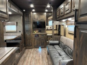 The interior of this Bloomer horse trailer features wood cabinetry, a leather sofa, a small dinette, wall-mounted TV, and laminate flooring. Windows with blinds line the walls, and recessed ceiling lights add warmth throughout the space. -Stillwater Trailer Sales