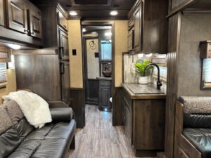 A modern Bloomer RV interior features dark wood cabinetry, a small kitchen with a plant on the counter, a black leather sofa with a white throw, and a view toward a bathroom with sink and mirror&mdash;perfect for your next 5 Horse adventure. -Stillwater Trailer Sales