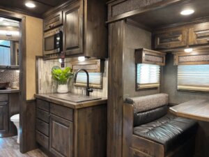 A compact Bloomer RV interior with dark wood cabinets, a small kitchen area featuring a sink and potted plant, a microwave, and a Signature dinette with cushioned seating; a bathroom is visible in the background. -Stillwater Trailer Sales