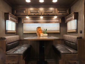 A dining area with two brown cushioned benches and a rectangular table, styled with an orange vase, plant, and candle. Surrounded by wooden cabinets and windows with blinds, it&rsquo;s a cozy space inside the 17' Signature Bloomer. -Stillwater Trailer Sales