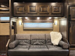 A Signature brown leather couch with patterned cushions and a white throw blanket sits against a wall with a window and closed blind. Above the couch are dark wooden cabinets, and the area is lit by three small lights. -Stillwater Trailer Sales