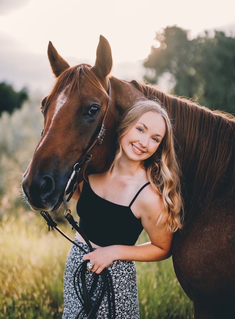 Hadley with long blonde hair in a black top and patterned skirt stands outdoors, smiling and leaning beside a brown horse in a grassy area with trees and sunlight in the background. -Stillwater Trailer Sales