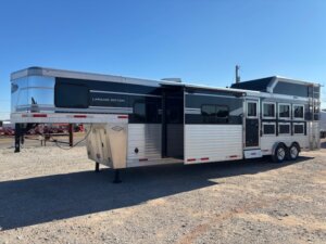 A large black and silver SMC 1706 gooseneck horse trailer with living quarters, labeled Laramie Edition, is parked on gravel under a clear blue sky. The 14' horse trailer features multiple windows and doors, two axles, and a rear ladder. -Stillwater Trailer Sales