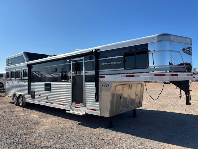 A large silver and black SMC Laramie Edition 4 Horse gooseneck trailer with multiple windows is parked on gravel under a clear blue sky. -Stillwater Trailer Sales