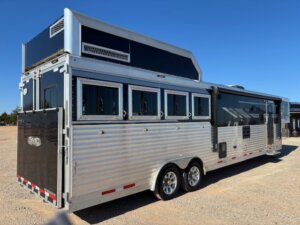 A large silver SMC 4 Horse gooseneck trailer with multiple side windows and a raised sleeping area is parked on a gravel lot under a clear blue sky. -Stillwater Trailer Sales