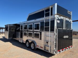 A large, silver and black SMC 4 Horse gooseneck trailer with multiple windows and vents is parked on gravel under a clear blue sky. It features three axles, a 14' slide-out, and the SMC logo along with "1706" on the back door. -Stillwater Trailer Sales