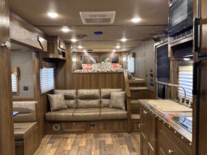 Interior of an SMC 4 Horse RV featuring wood paneling, a brown leather sofa, a kitchen area with sink and stove, and a raised sleeping area with bedding and pillows. Steps lead up to the bed; blinds cover the windows. Model: 1706. -Stillwater Trailer Sales