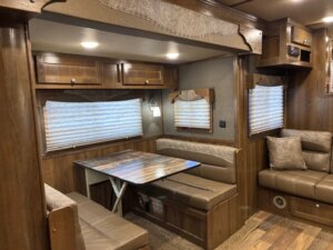 A dining area inside a 14' horse trailer features a wooden table, two bench seats, and built-in cabinets above. A sofa with cushions is on the right, and windows with pleated blinds provide natural light. -Stillwater Trailer Sales