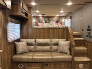 A tan leather couch with two patterned pillows sits in a wood-paneled SMC 4 Horse interior. Stairs on the right lead to a lofted bed with a leopard print blanket and pillows in this stylish 14' horse trailer. Windows are covered with blinds. -Stillwater Trailer Sales