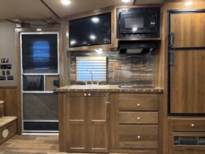 A compact RV kitchen in a 14' horse trailer with wood cabinets, small sink, cooktop, microwave, refrigerator, and a TV mounted above the counter. A window with shade sits over the sink, and a door is located to the left. Model: SMC 4 Horse 1706. -Stillwater Trailer Sales