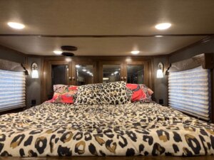 A bed with leopard print bedding is centered in a compact SMC 1706, 14' horse trailer, featuring wood-paneled cabinets and mirrored doors at the head. Two windows with blinds and wall lamps flank the bed, creating a cozy retreat in this SMC 4 Horse setup. -Stillwater Trailer Sales