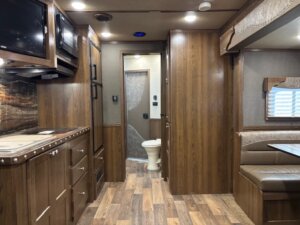 Interior of a 14' horse trailer with wood paneling, a kitchenette on the left, dining area on the right, and a rear door leading to a small bathroom with visible toilet. The floor features wood-patterned vinyl for easy cleaning. -Stillwater Trailer Sales