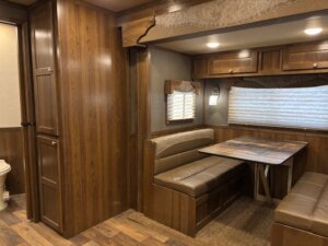 Interior of an SMC 4 Horse Trailer featuring a wood-paneled dining area with booth-style table and benches, overhead cabinets, carpeted and wood flooring, plus a partial view of the bathroom with a toilet. -Stillwater Trailer Sales