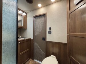A compact bathroom inside a 4 horse trailer features wood paneling, a small sink with a mirror above, a closed wooden cabinet, frosted glass partition, toilet, and a decorative interior door with control switches on the wall. -Stillwater Trailer Sales