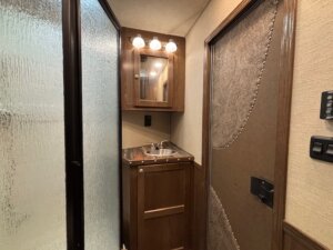 A small bathroom area in a 4 horse trailer features a wooden vanity, round sink, mirrored cabinet above with three lights, a patterned door, and a frosted glass shower enclosure. -Stillwater Trailer Sales