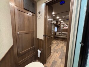 View from a bathroom inside a 14' trailer RV, showing wood-paneled walls, a toilet, a cabinet, and toilet paper holder. Through the doorway, a hallway leads to a living area with a brown sofa and overhead storage. -Stillwater Trailer Sales