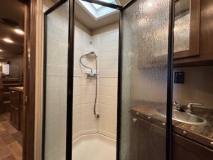 Corner shower with frosted sliding glass doors in the SMC 1706 4 Horse RV bathroom; white tiled shower walls, handheld showerhead, skylight above, and a compact sink with faucet and countertop to the right. -Stillwater Trailer Sales