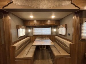 The image shows the dining area of an SMC 4 Horse RV, featuring two long cushioned benches facing each other, a rectangular wooden table in between, wooden cabinets above, and windows with blinds on three sides. -Stillwater Trailer Sales