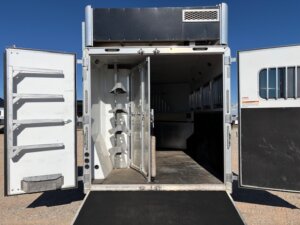 An SMC 4 Horse trailer stands with both rear doors open, revealing an empty 14' trailer interior with saddle racks on the left, partitioned stalls on the right, and a lowered ramp on gravel beneath a clear blue sky. -Stillwater Trailer Sales