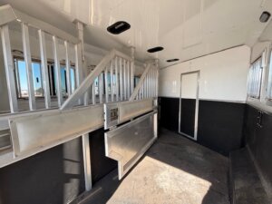 Interior of a 4 horse trailer with metal partitions, black lower walls, white upper walls and ceiling, barred windows, and an open door at the far end. Sunlight streams in, casting shadows on the floor of this spacious SMC horse trailer. -Stillwater Trailer Sales