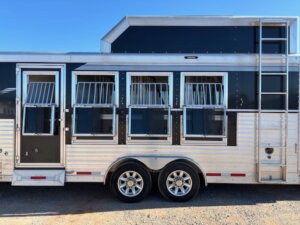 A side view of a silver and black SMC 4 Horse trailer with four barred windows, two axles with shiny wheels, a ladder on the right, and a clear blue sky in the background. Model 1706. -Stillwater Trailer Sales