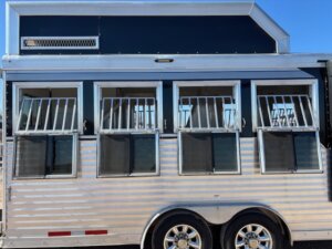 A close-up side view of a silver SMC horse trailer with barred windows and black accents. The 4 horse trailer has two visible wheels and is parked under a clear blue sky. -Stillwater Trailer Sales