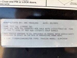 A close-up of a metal label on a 14' SMC 4 Horse trailer showing manufacturing details, weight limits, tire and rim specs, compliance notice, VIN, trailer type 1706, and model number. The label is slightly worn. -Stillwater Trailer Sales