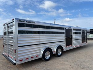 Silver aluminum Elite 30' livestock trailer with two axles is parked on a gravel lot under a partly cloudy sky. Perfect for show cattle, the trailer features horizontal slats, tinted windows, and Elite Trailers branding on the upper left. -Stillwater Trailer Sales