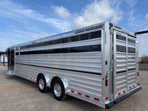 A large silver Elite 30 livestock trailer with two axles, ribbed metal sides, tinted windows, and red and white reflective tape is parked on gravel under a partly cloudy sky—perfect for hauling show cattle or as a versatile Cattle Combo. -Stillwater Trailer Sales