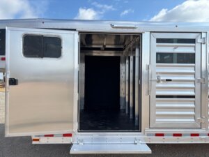 A silver Elite Combo livestock trailer with an open side door reveals a dark, empty interior lined with dividers. The metallic exterior features ventilation slats and a window, while the sky and ground can be seen in the background. Perfect for show cattle. -Stillwater Trailer Sales