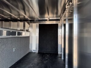 Interior view of an empty Elite 30 metal trailer with exposed framework, a carpeted panel on the left, hooks along the right wall—ideal for show cattle—and a large black door at the far end. The floor is clean and the space is well-lit. -Stillwater Trailer Sales