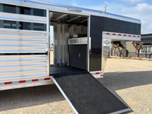 A silver Cattle Combo livestock trailer with a black ramp extended to the ground, showing an open door and interior compartments, is parked on a gravel lot under a partly cloudy sky—the perfect choice for hauling show cattle. -Stillwater Trailer Sales