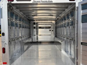The image shows the interior of an empty Elite 30 aluminum livestock trailer with vented side panels, a textured metal floor, and a partition gate at the far end. Perfect for hauling show cattle, the trailer is clean and well-lit by natural daylight. -Stillwater Trailer Sales